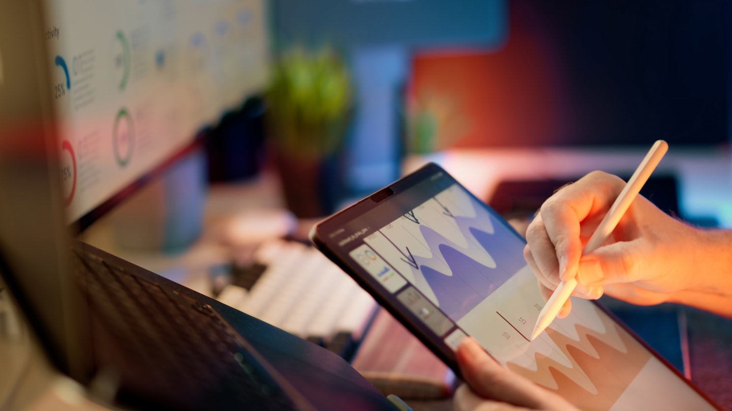 Hands use a stylus on a tablet displaying data charts.
